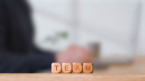 Business-Concept-Wooden-Letter-Cubes-Or-Dice-Spelling-Team-With-Office-Person-Using-Mobile-Phone-In-Background